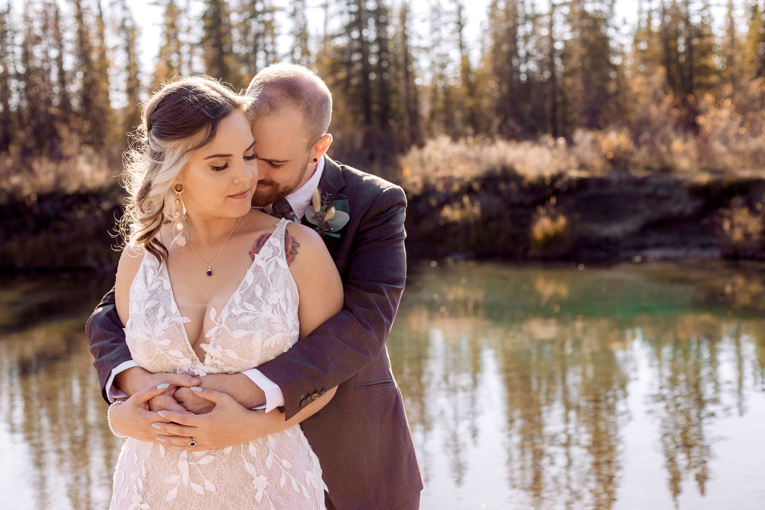 Groom wrapping his arms around bride from behind with his nose nuzzled into new neck. Bride looks down gently to the side. The couple is in front of a river with spruce trees in the background.