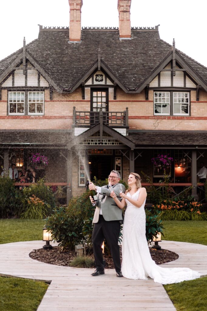 Calgary wedding photographer photograph of a bride and groom doing a champagne pop in front of the Bow Valley Ranche Restaurant