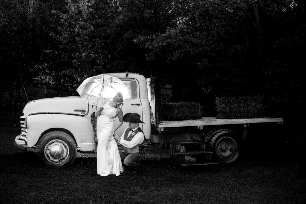 Groom helps bride with her bustle in the rain in front of an old truck at The Woven Willow