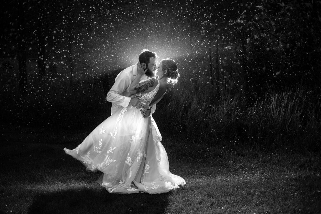 Groom slight dips his bride and leans in for a kiss while a flash illuminates the rain drops
